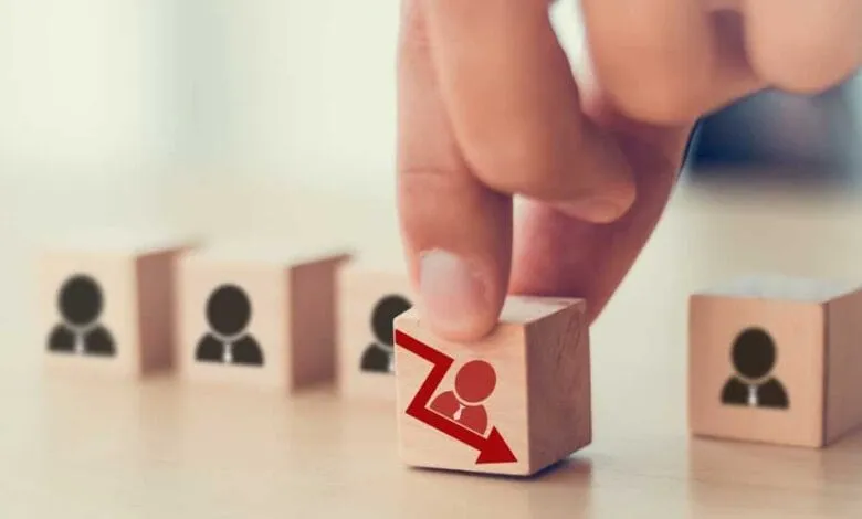 Hand placing wooden block with downward red arrow and person icon, symbolizing job loss or layoffs.