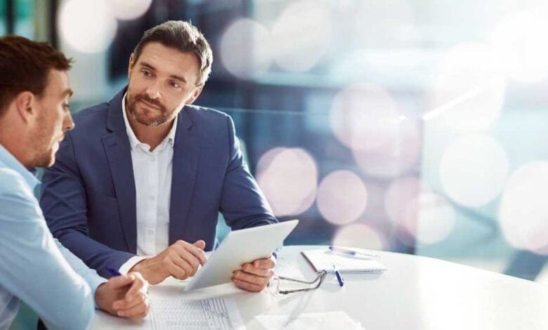 Two businessmen sit at a table, reviewing documents and a tablet. One man gestures towards the screen.