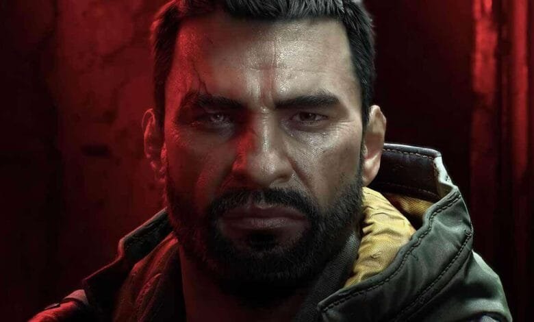 Close-up of a man with a serious expression, dark beard, and scar, illuminated by red and dim light.