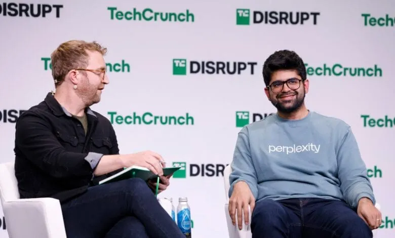 Two men sit on stage at a TechCrunch Disrupt event. One takes notes, the other smiles, wearing a Perplexity AI sweatshirt.