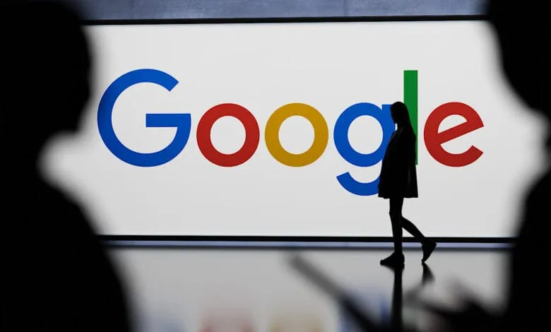 Silhouette of a person walking past a large Google logo displayed on a bright white screen.