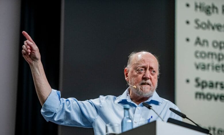 Older man with beard giving presentation, gesturing with hand.