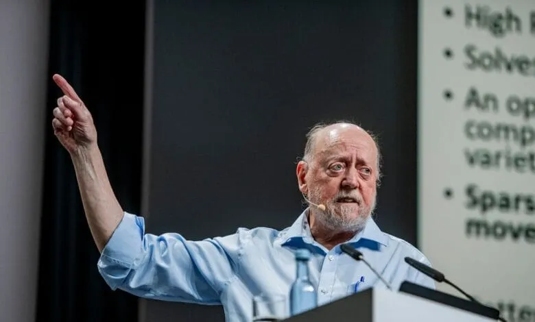Older man with beard giving presentation, gesturing with hand.