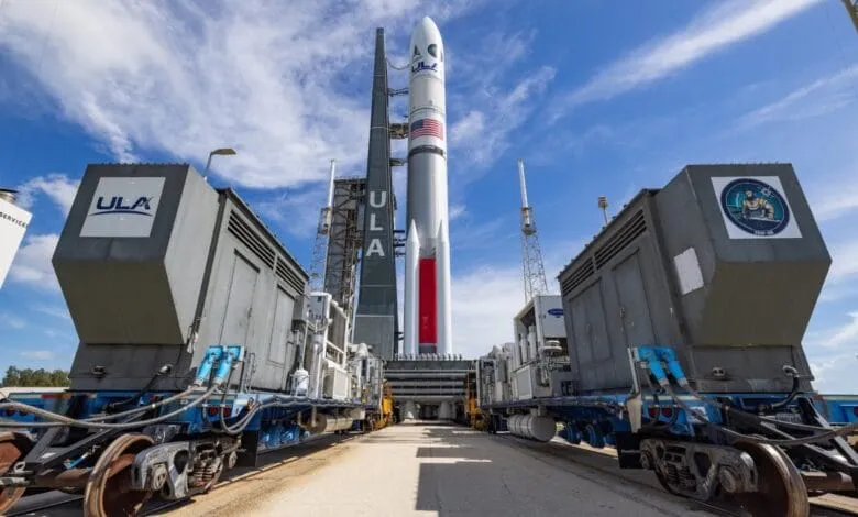 A large rocket stands tall on a launchpad, flanked by support equipment on railway cars under a bright blue sky.