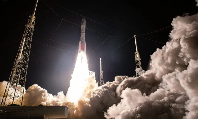 A rocket launches at night, surrounded by bright flames and smoke, with tall metal structures visible in the background.