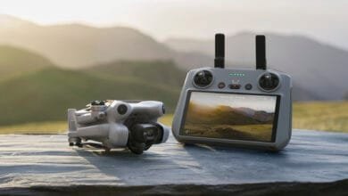 A small drone and its controller sit on a dark gray surface against a blurred background of rolling green hills at sunset.
