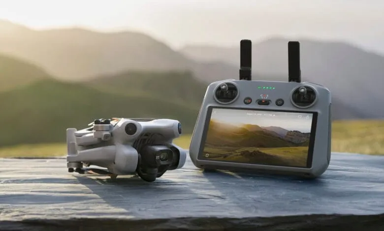 A small drone and its controller sit on a dark gray surface against a blurred background of rolling green hills at sunset.
