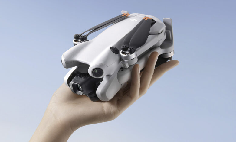 A hand holds a small, white DJI Mini 3 Pro drone with orange accents. The drone's propellers are folded.