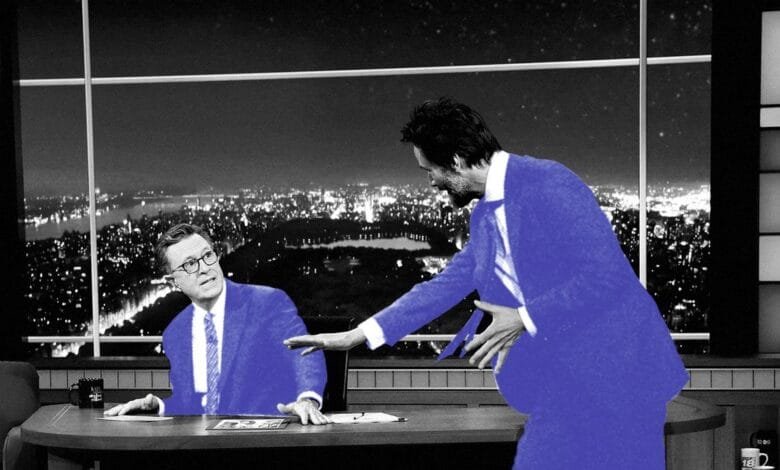 Stephen Colbert, in a blue suit, sits at a desk as a man in a blue suit approaches him. Nighttime city view is visible through a large window.