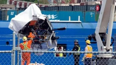 Damaged submersible being lifted by a crane onto a ship, workers in safety gear oversee the operation.