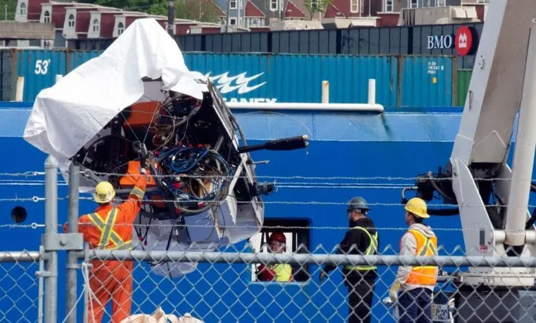 Damaged submersible being lifted by a crane onto a ship, workers in safety gear oversee the operation.