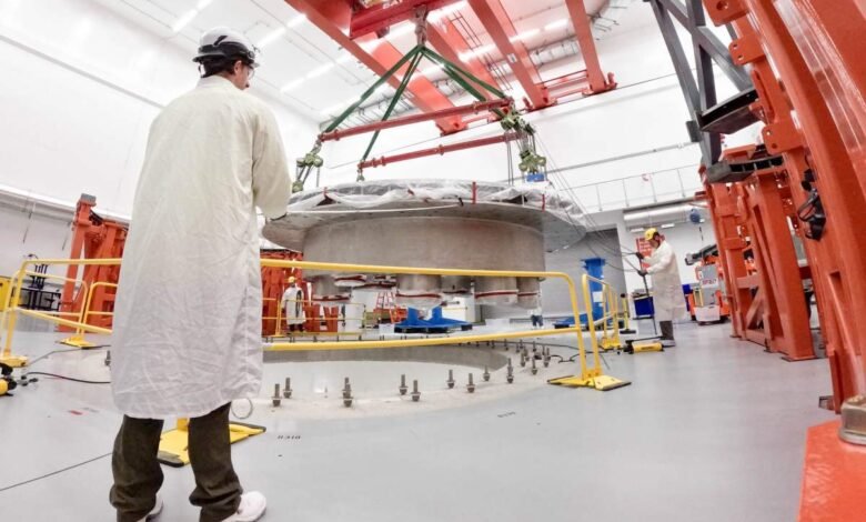 Scientists in lab coats oversee the precise installation of a large, circular cryostat base using an overhead crane.