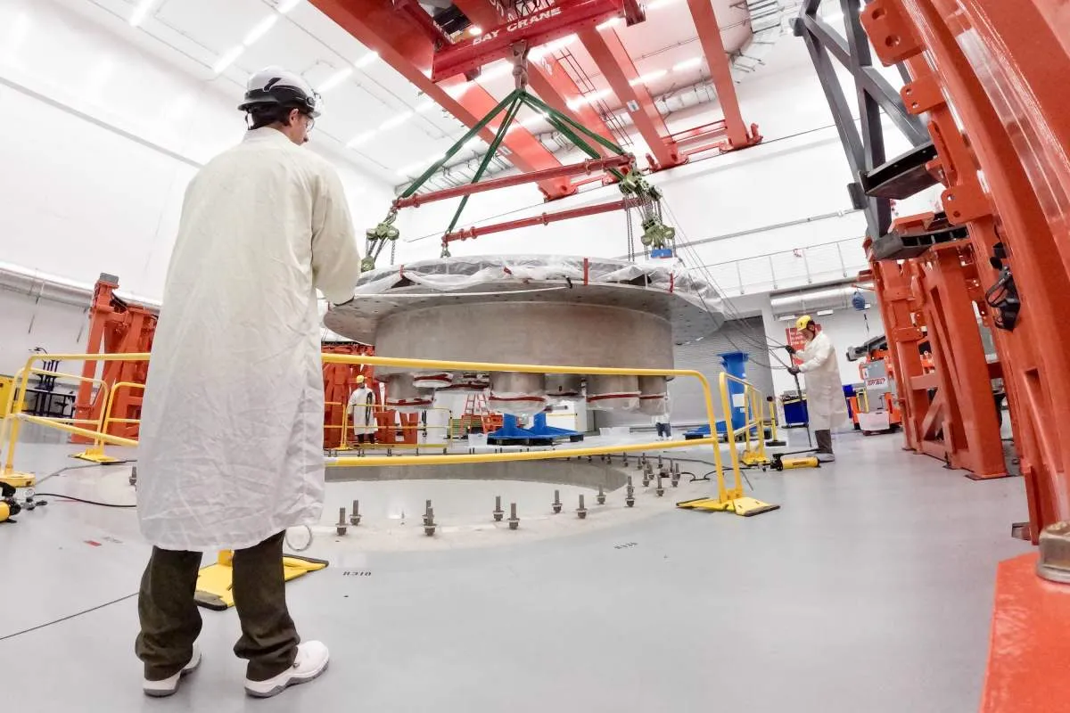 Scientists in lab coats oversee the precise installation of a large, circular cryostat base using an overhead crane.