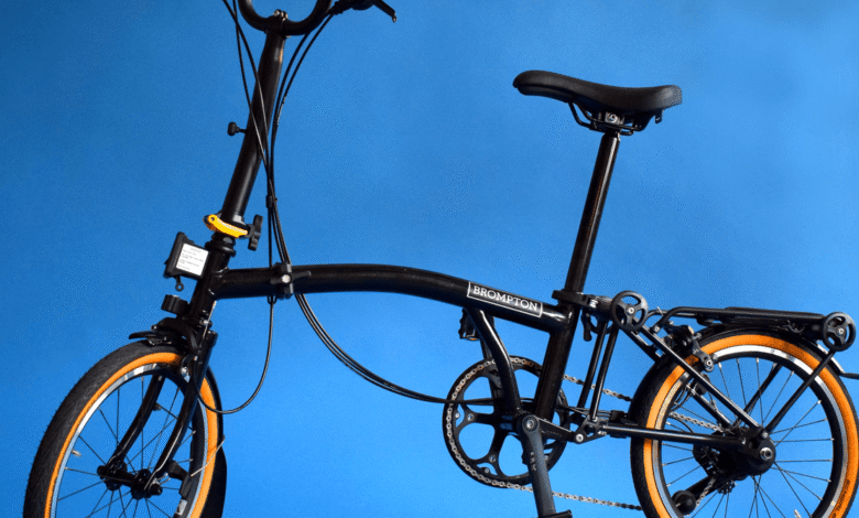 Black Brompton folding bicycle with orange-rimmed wheels against a blue background.