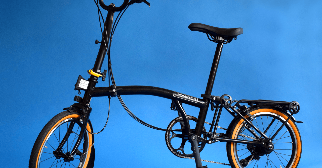 Black Brompton folding bicycle with orange-rimmed wheels against a blue background.
