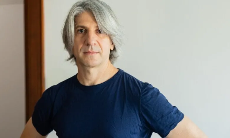 Headshot of a man with graying hair, wearing a dark blue t-shirt, against a light background.