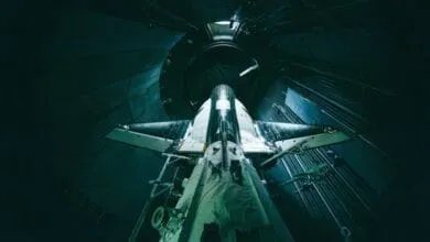 A spacecraft, possibly Dream Chaser, is shown inside a dark hangar, viewed from below. Its sleek design and dark teal color are prominent.