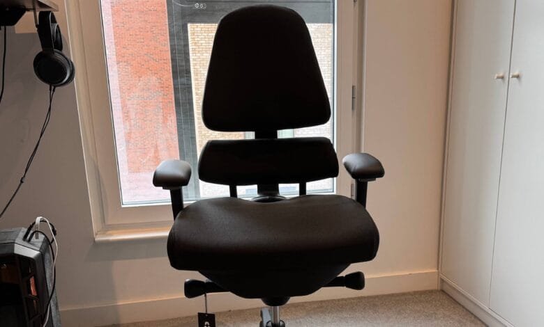 Black ergonomic office chair sits near a window, with headphones and a computer case visible in the background.