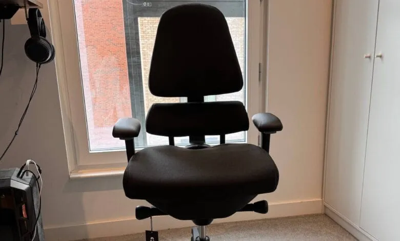 Black ergonomic office chair sits near a window, with headphones and a computer case visible in the background.