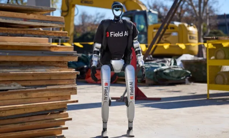 A bipedal robot, wearing a Field AI shirt, stands on a construction site.
