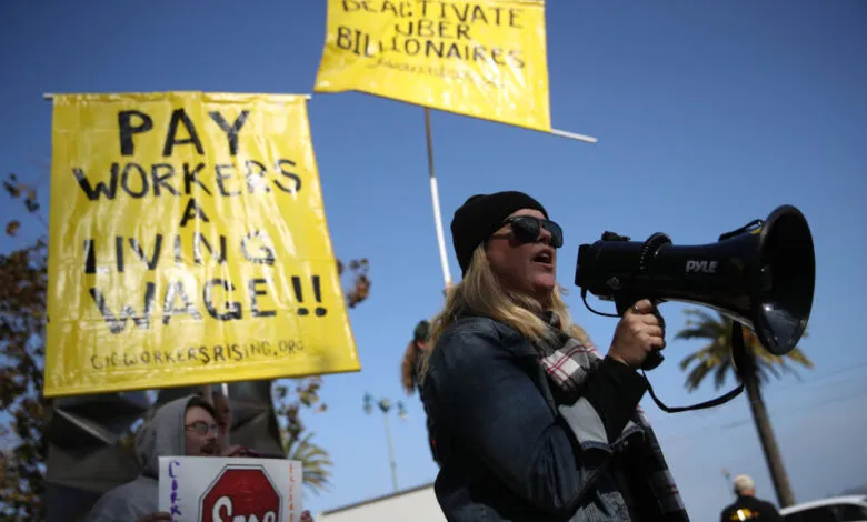 A woman uses a megaphone at a protest, demanding fair pay for Uber and Lyft drivers. Yellow signs read 'Pay Workers a Living Wage!'