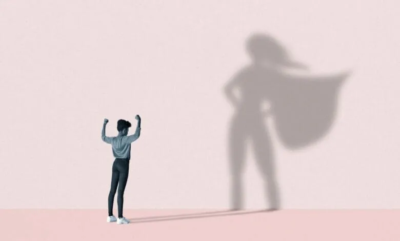 A woman stands with arms raised, her shadow cast as a superhero figure with a cape.