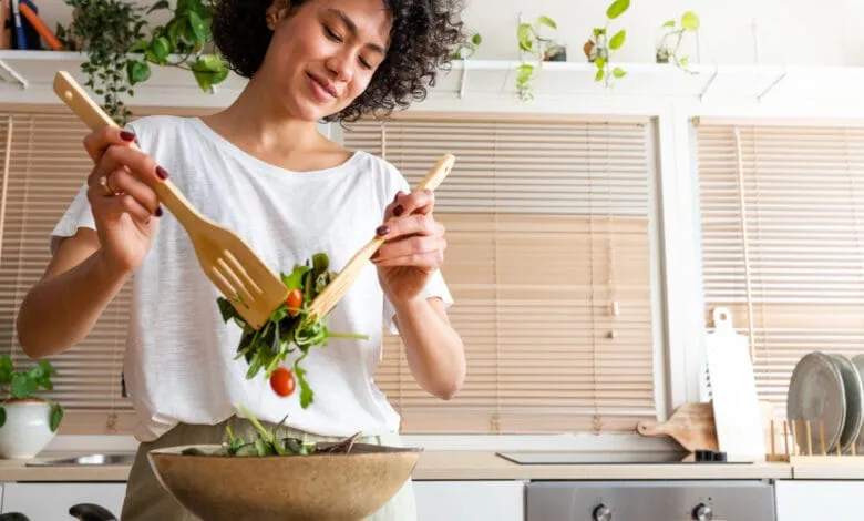 A woman with curly hair joyfully tosses salad greens and cherry tomatoes into a bowl with wooden utensils.