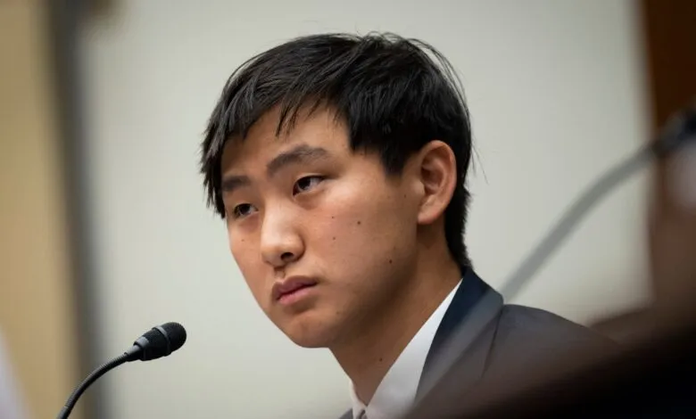 Close-up of a young man with dark hair, wearing a suit, seated at a microphone.
