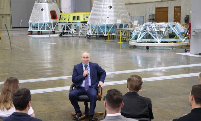 Russian President Vladimir Putin speaks to a group in a large industrial hangar with spacecraft components.