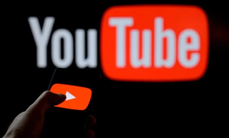 Close-up of a hand holding a smartphone displaying the YouTube play button, with a blurred YouTube logo in the background.