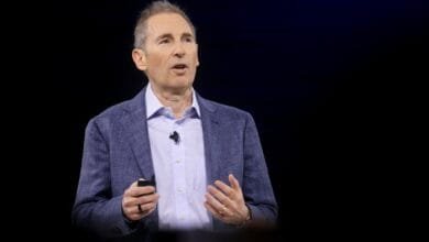 A man in a blue blazer speaks at a conference, holding a small device.