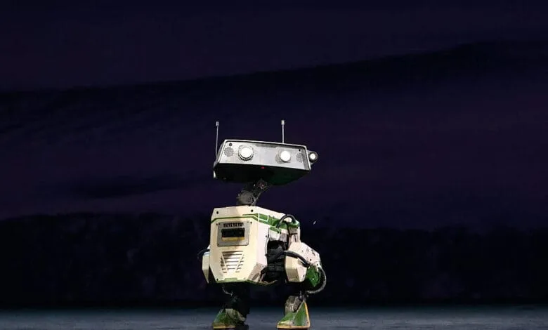 A humanoid robot, primarily off-white and green, stands on a dark stage. It has a boxy design with lights on its head.