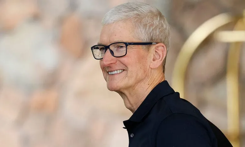 Tim Cook, Apple CEO, smiles subtly while wearing glasses and a dark polo shirt.
