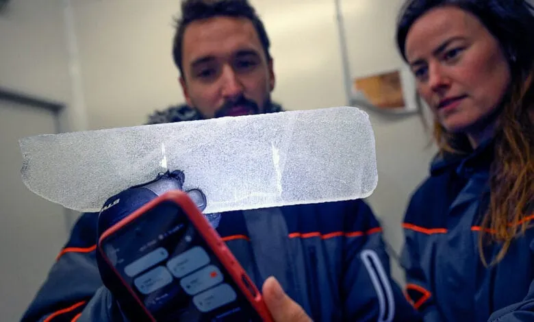 Two scientists examine a translucent ice core sample, backlit by a smartphone, revealing internal structures. Antarctic climate research.