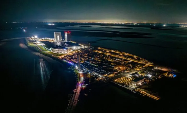 Aerial nighttime view of a coastal city with illuminated buildings and parking lots.