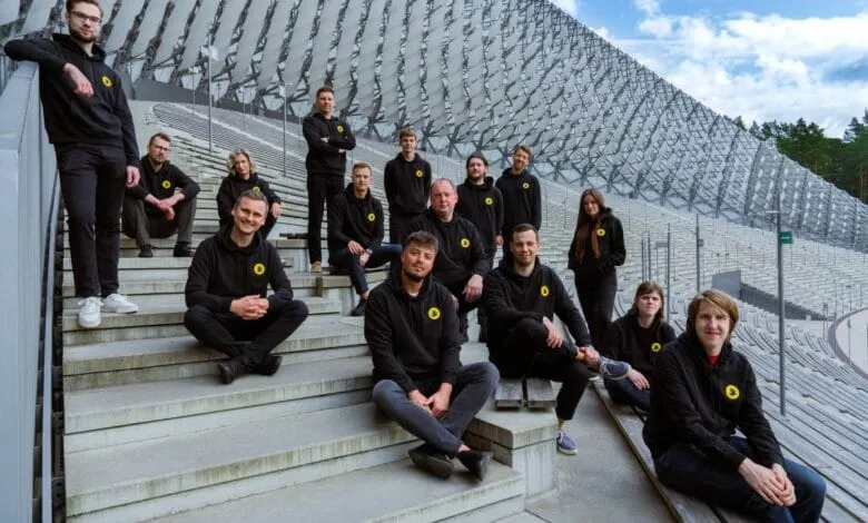 Fifteen people in black hoodies with a yellow logo pose on stadium steps.