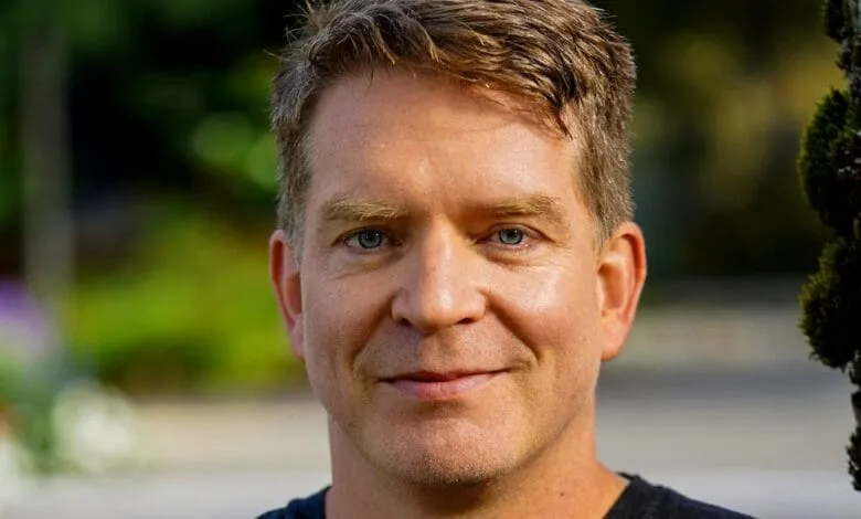 Close-up portrait of a man with light brown hair and blue eyes, smiling gently. He's outdoors, near a tree.