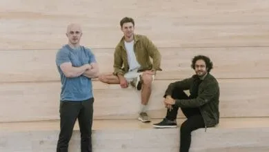 Three men stand and sit against a light wood wall. One stands with arms crossed, two sit with legs crossed.