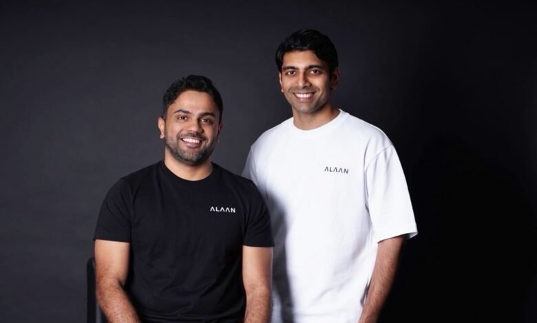 Two men, smiling, wear black and white ALAAN brand t-shirts against a dark background.