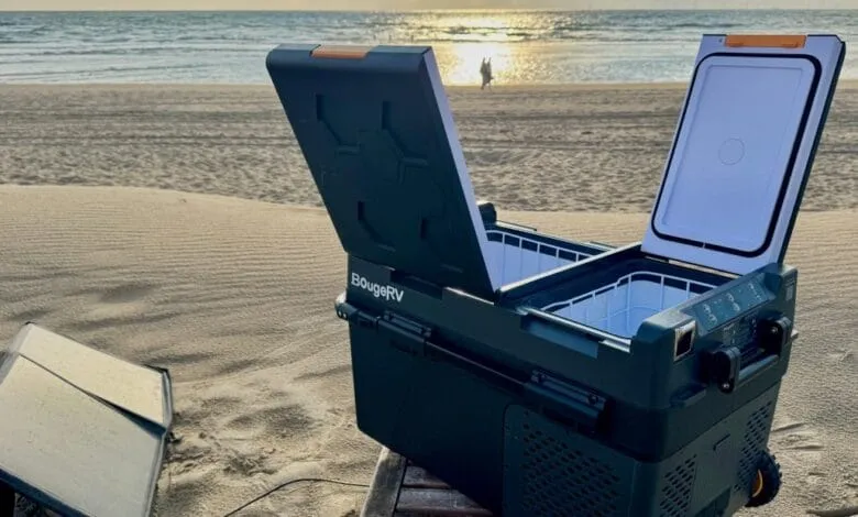 A BougeRV portable fridge freezer sits open on a sandy beach with a solar panel nearby, ocean and sunset in the background.