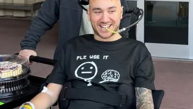Smiling man in wheelchair with medical tube, wearing a shirt that says 'Plz use it', with a cake nearby.