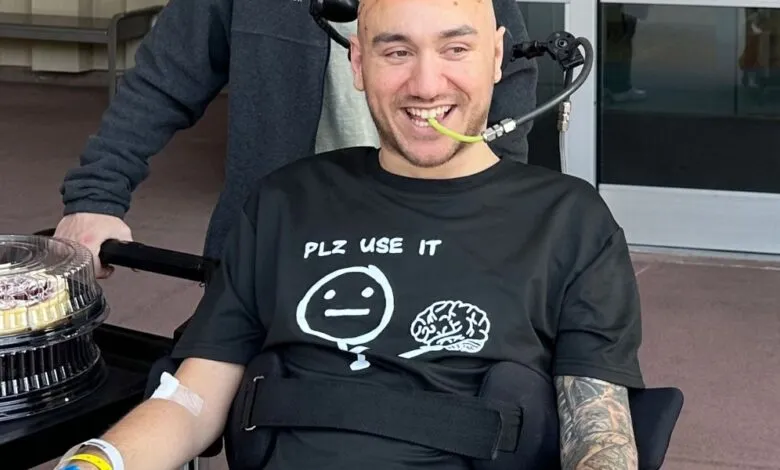 Smiling man in wheelchair with medical tube, wearing a shirt that says 'Plz use it', with a cake nearby.