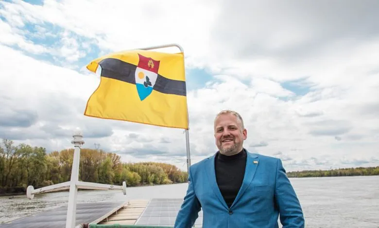 Vít Jedlička, President of Liberland, stands on a boat with the Liberland flag waving behind him.