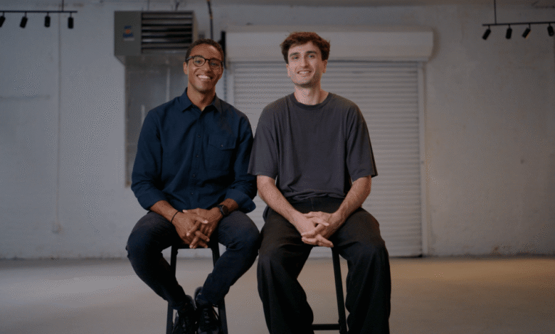Two men smiling, seated on stools in a minimalist studio setting.