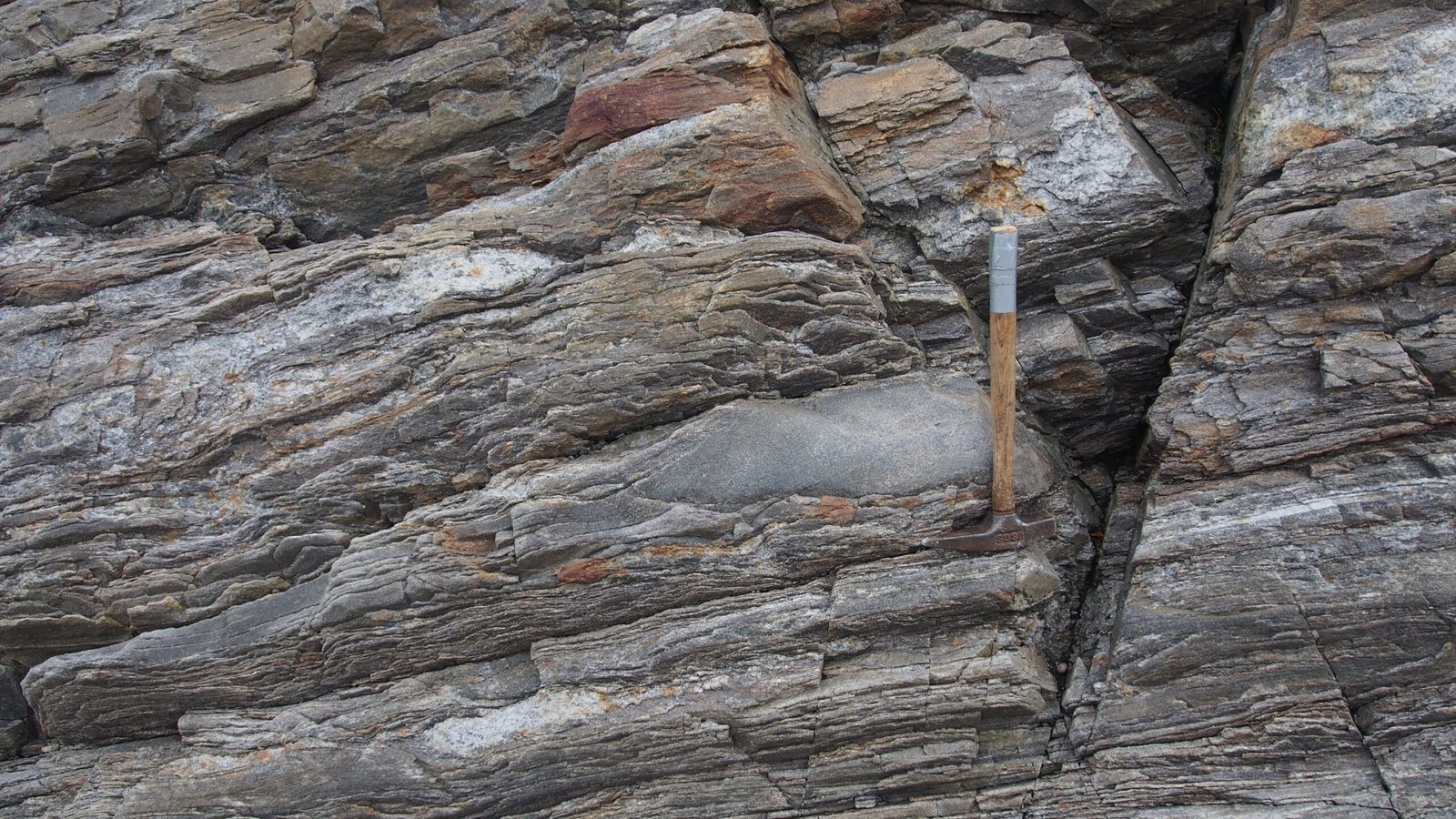 Close-up of layered rock formation with a rock hammer for scale, showing variations in color and texture.