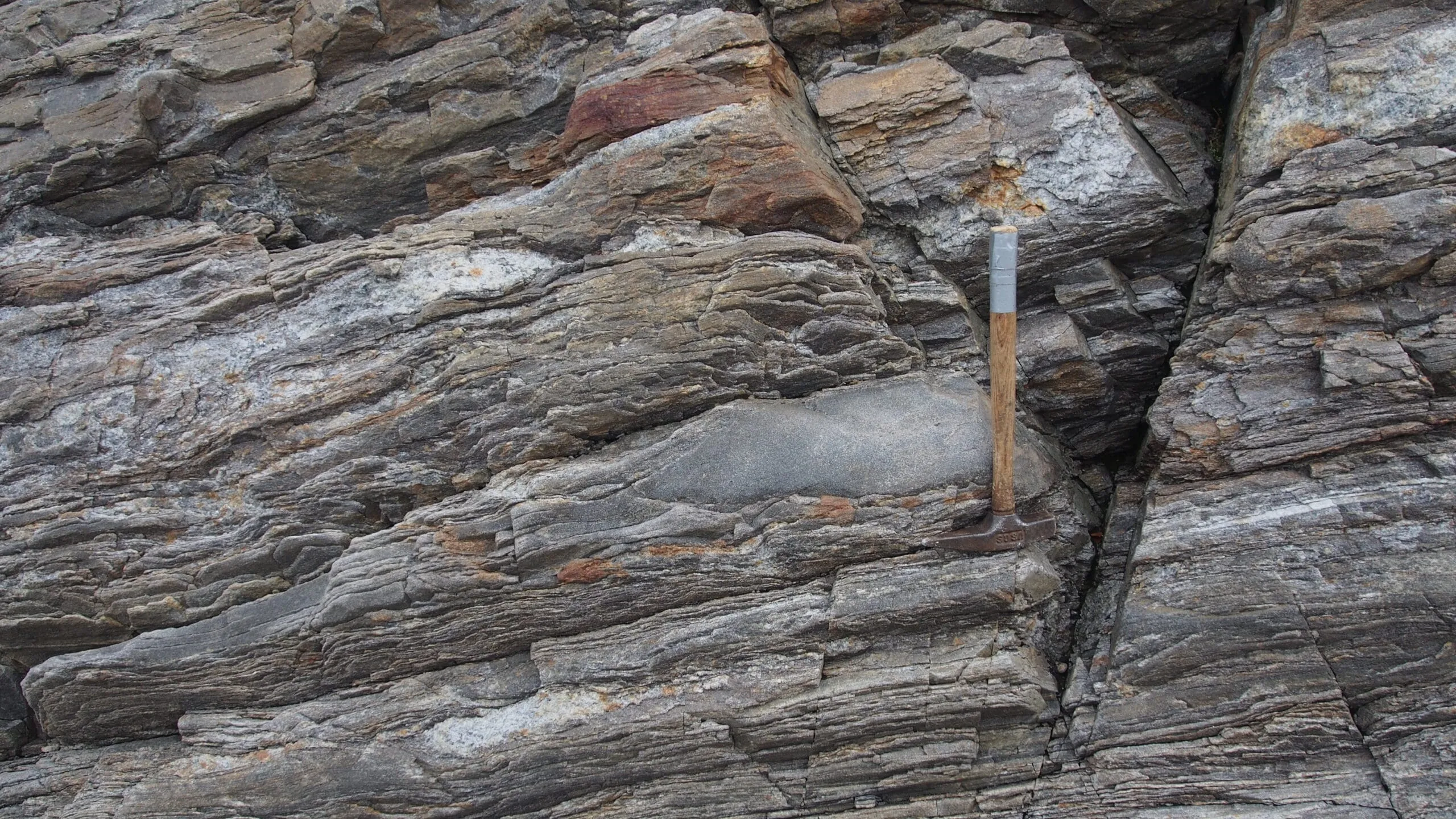 Close-up of layered rock formation with a rock hammer for scale, showing variations in color and texture.