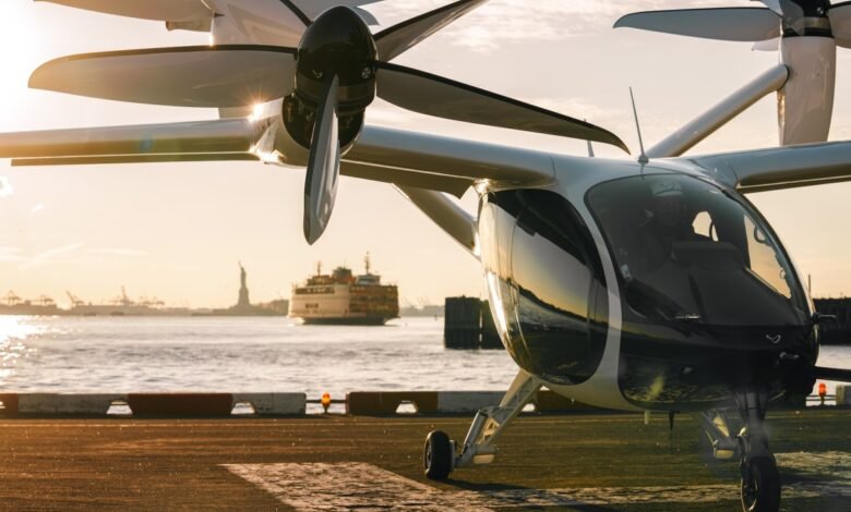 A white electric air taxi sits on a tarmac at sunset, with the Statue of Liberty visible in the background.