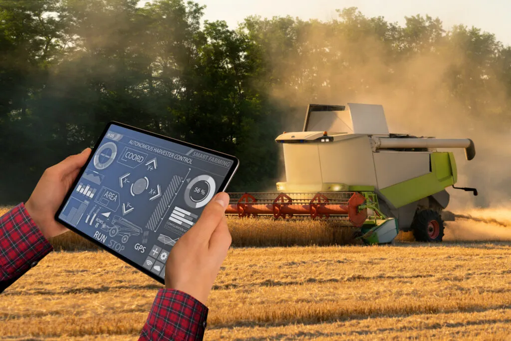 Farmer uses tablet to monitor autonomous harvester in field. GPS data and harvest progress shown on screen.