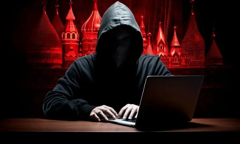 A hooded figure sits at a desk in a dimly lit room, working on a laptop with a red-lit backdrop of Kremlin-like buildings.