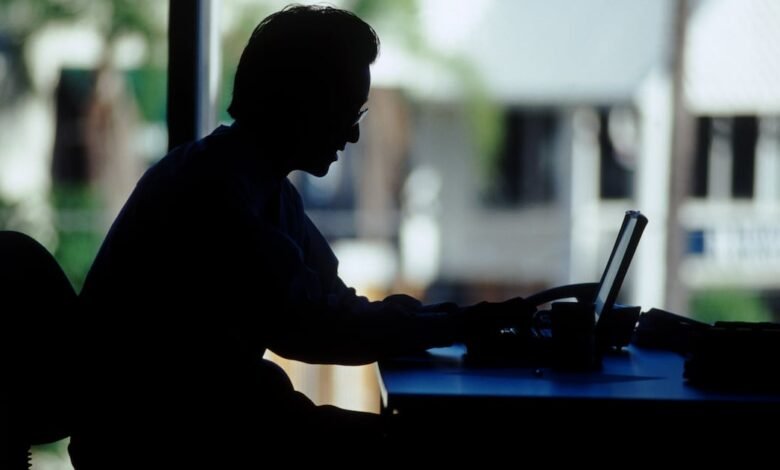 Silhouette of a person working on a laptop at a desk, near a window with a blurred outdoor background.